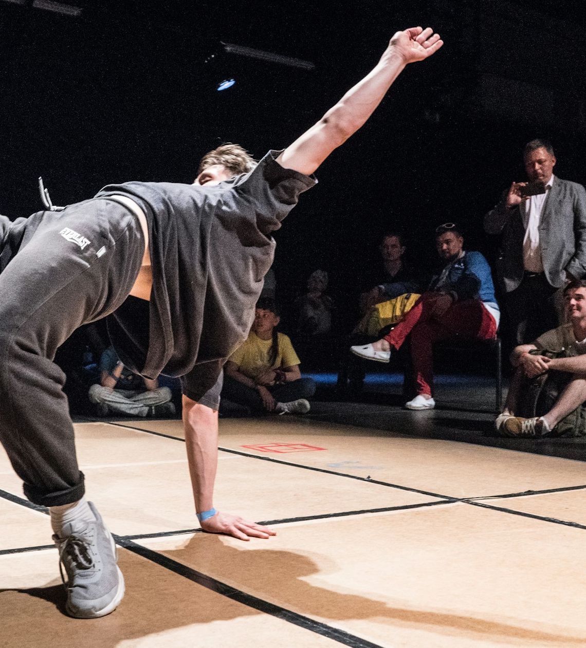 A b-boy mid freeze on the floor with the crowd watching from the dark
